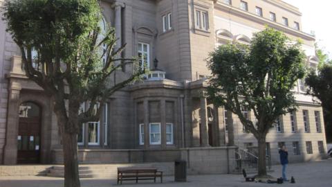 The Royal Court in St Helier is a grand old stone building which has columns, arches, different shaped windows and a large wooden entrance door. In front of it there are two trees, a wooden bench and a bin. A man is standing under one of the trees.