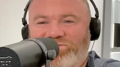 A man, Wayne Rooney, has a beard and is wearing headphones and talking into a microphone
