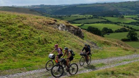 Three cyclists are riding uphill on a small stone path. Surrounding them are rolling green hills that stretch into the horizon. The sky is bright with lots of white cloud.