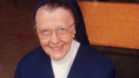 An archive colour photograph of Sister Agnes. She is wearing glasses, a blue dress and white top. She is smiling at the camera.