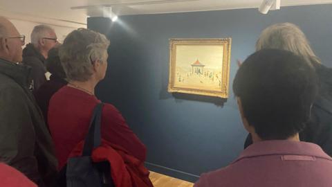 A group of people standing round a painting by LS Lowry which depicts a beach and people playing around a bandstand 