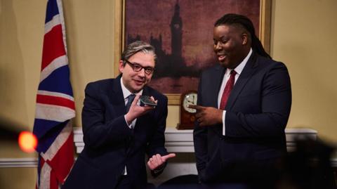 Two actors in a set resembling 10 Downing Street - one is appearing as Keir Starmer, the other as David Lammy, both wearing suits. A union jack and painting of Big Ben are behind them