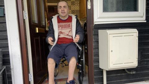 A man has dark, close-shaven hair and is wearing a grey hoodie, a red T-shirt and blue shorts. He is using a wheelchair and is in the doorway of a flat with a step outside.