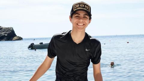 Zak is stood on a beach with the sea behind him. He is smiling at the camera. The sea is a clear blue and it is a sunny day.