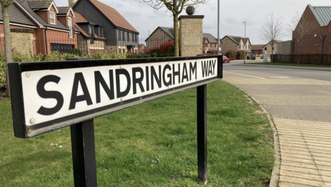 A street sign for Sandringham Way with a housing estate and railings visible in the background.