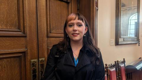 lilac has long dark hair, with a pink fringe, dangly earrings, an eyebrow piercing. She is wearing a sparkly necklace and smart jacket. She is standing in the council chamber, smiling.