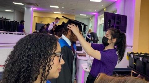 Students being fitted with gowns and mortarboards for graduation.  There is one woman in the foreground with long dark hair and glasses, and a man with short dark hair and beard being fitted with a mortarboard by a woman with long dark hair wearing a purple T-shirt and a mask.