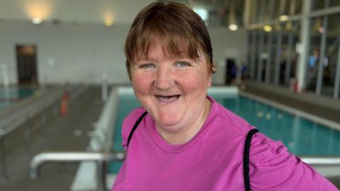 Amy Mercer is wearing a pink long sleeved t-shirt and smiling at the camera. She has short light brown hair and blue eyes and is standing in front of the swimming pool where her class is held.