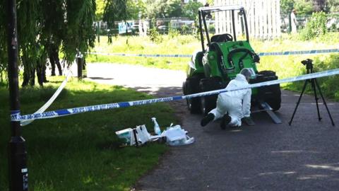 A person in white overalls is on the ground at the front of a small, green, single-seater, four-wheeled vehicle which has two forks at the front. It has a roof over the driver's seat.  It is cordoned-off on a path, and has blue and white police tape around trees.