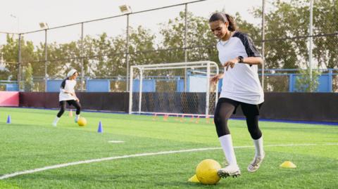 Two teenage girls dribbling yellow footballs around cones on an Astro Turf pitch. They are wearing kits made of white shirts and black leggings. They are playing on a five-a-side pitch surrounded by fencing.
