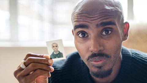 Sir Mo Farah pictured looking into the camera while holding a passport photo of himself as a child