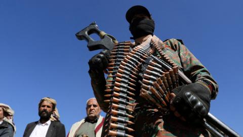 A Houthi policeman stands in front of a camera with a gun