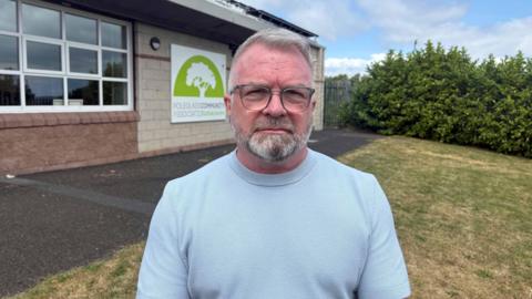 A man with white hair and glasses looks at the camera. He is wearing a pale blue t-shirt. He is standing outside on a patch of grass next to a building. The building has a sign on it which reads: 'POLEGLASS COMMUNITY ASSOCIATION'.