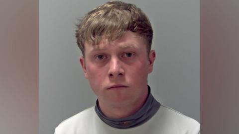A police custody photo of Levi Williams. He has light brown hair which over hangs on to his forehead. He wears a white top with a grey turtleneck. 