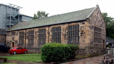 This image features an old stone building with a gabled roof and large rectangular windows that have a grid-like design. The structure is made from stone or brick blocks in varying shades of brown and grey, giving it a weathered, traditional appearance. In front of the building, there is a small grassy area with a bush.
To the left of the building, a red car is parked near the entrance. The surroundings include paved walkways and, in the background, some modern buildings.
