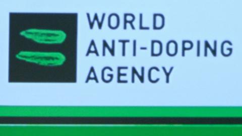 The World Anti-Doping Agency logo (a black square with two green streaks through it) next to the name of the agency in black on a white screen at a conference