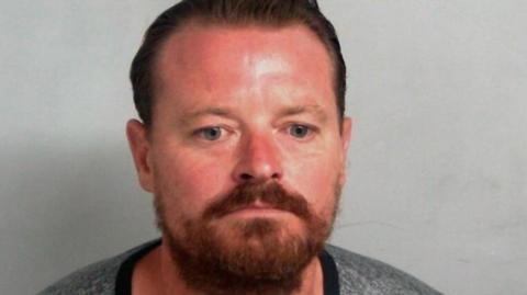 Lee Gower pictured wearing a grey T-shirt in police custody. He has brown hair and a beard.