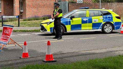 Police officer standing behind cordon on a road with police car behind and homes in the background.