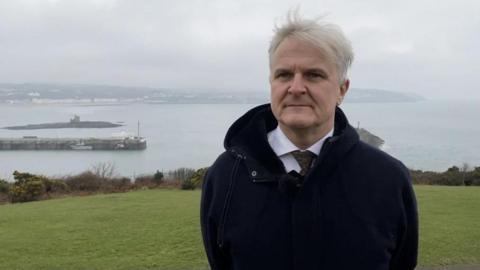 Diccen Sargent standing on the green hillside on Douglas Head with Douglas Bay in the background. He has grey hair and is wearing a white shirt and green tie, with a black coat.