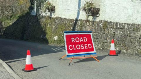 A red road closed sign on a rural road with traffic cones alongside it, and a white wall behind