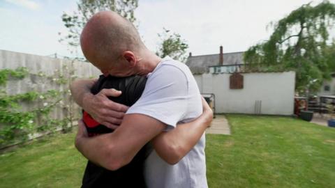 Gavin Bowden and Oscar are hugging each other in a garden. Gavin has his eyes closed and is smiling and he has his hands on Oscar's back and Oscar's hand is on Gavin's back.