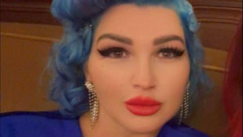 Tania Williams is inside looking to the camera. She has wavy blue hair and make-up on, including bright red lipstick and black mascara. She is wearing a dark blue dress.