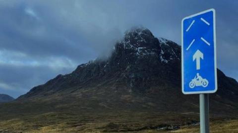 The sign is a long rectangle shape. It is blue with white markings, including an arrow point skywards, and a motorbike and rider. In the background is the mountain Buachaille Etive Mor. The sky is dark and cloudy.