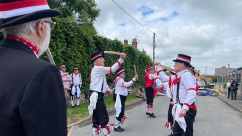 A Morris dancing troupe waves their batons in the air, dressed in traditional clothing. Bystanders watch the dancing on the village street, which is decorated in Union Jack bunting.