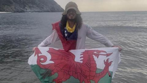 Yn y llun yma mae Ollie yn cario baner Cymru ar lan y mor. Mae e'n gwisgo siwmper hufen a chap hufen hefyd. Mae ganddo wallt brown hir.