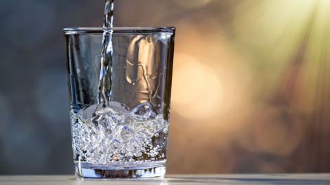 A galss being filled with water. There are rays of sunlight behind.