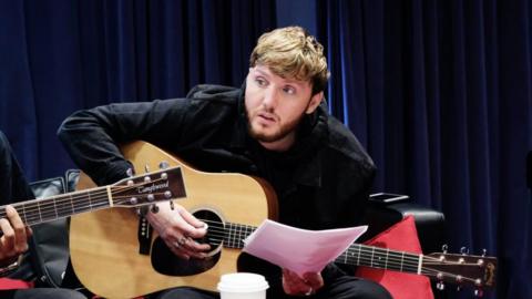 Man with dark hair and short beard playing a guitar