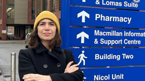 A woman wearing a black coat and yellow beanie hat stands in front of the Stratford Hospital sign. The sign includes directions and names of the various hospital departments and units.