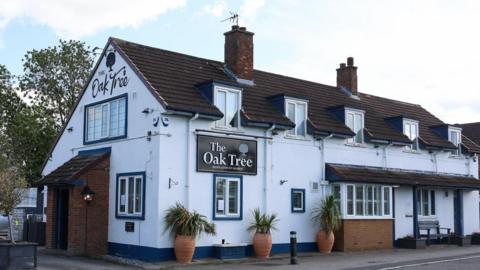 The exterior of The Oak Tree inn. It is a large, two-storey white building white with potted plants outside. The first floor has a row of five dormer windows and there are entrances at the front and side. The pubs name is painted in a script style writing on the side, plus there is a more traditional black sign on the front also containing the pubs name.