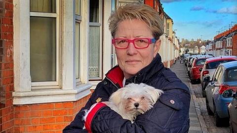 Woman with medium-length light brown hair and glasses wearing a blue coat holds a light-coloured terrier