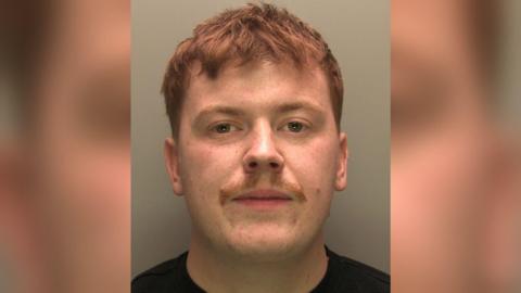 A police custody image of Brandon Barker. He has short, cropped red hair, freckled skin and a small moustache. He is staring into the camera and is wearing a black t-shirt.