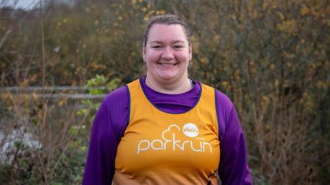 Alannah Mathewson is a woman in her 20s wearing a purple top and an orange vest with the Parkrun logo in white. She is standing in front of some trees with a river behind
