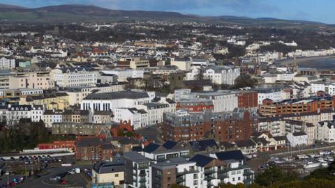 A panoramic shot of Douglas with houses, shops and apartments seen on street level. The sea is to the right, whilst hills can be seen in the distance.