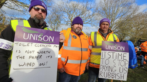 Terry Money, wearing sunglasses and a Unison hat, holds up a sign that says Unison: Toilet cleaners pay is not worth the paper it's wiped on. He stands next to Jim Kirkwood, dressed in a hi-vis orange jacket. Next to him stands Phil Hammond. He is wearing a hi-vis yellow jacket and a Unison hat and holds a sign that reads Unison: It's time to stop paying us rubbish