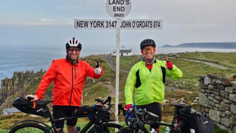 Mark and Ian standing with their bikes in front of the landmark sign at Land's End, Cornwall.