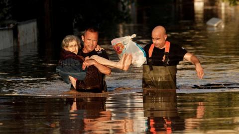 Greek firemen carry an elderly woman through floodwater