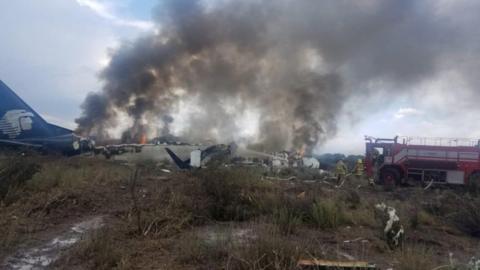 Firefighters douse a fire as smoke billows above the site where an Aeromexico passenger jet crashed