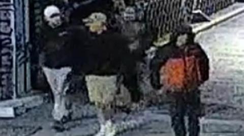 A grainy CCTV screenshot that shows four men walking over a bridge. It is hard to make out any details but one wears a black and red jacket with the hood up