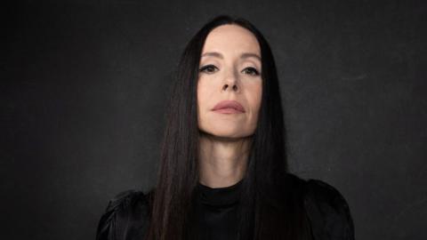 A woman with long straight black hair, wearing a black top, is standing in front a black backdrop. She is looking into the camera.