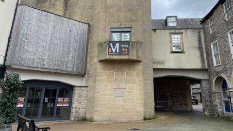 The picture shows the outside of the Amulet Theatre in Shepton Mallet. There are stone walls and wooden panels.  There is a bench to the left of the picture,