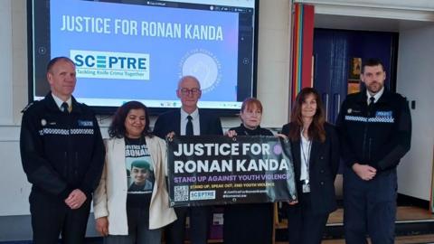A group of people standing infront of a class of students holding a sign about knife crime and justice for Ronan kanda