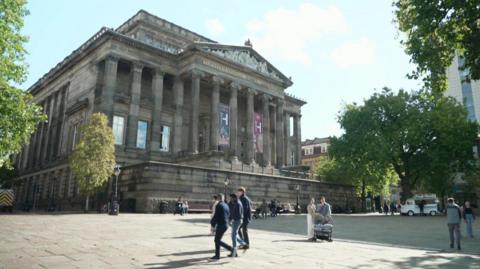 The Harris Museum front exterior with classical columns rises above a public square with people walking across. Trees line the sides of the square.