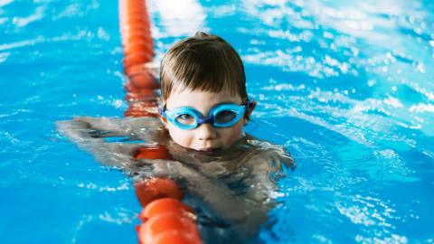 a boy in a swimming pool wearing swimming goggles holds onto a floating pool divider while staring at the camera
