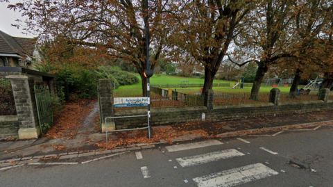 A large park is in the background, behind a row of trees and a playground. The sign by its entrance says Ashcombe Park and it is on a gate in front of a road with a zebra crossing.