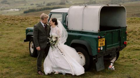 A newly-married couple pose in front of a dark green Land Rover Defender that is parked on a grassy hill. The bride wears a floor-length white gown with lace details and a short veil. She holds a bouquet of white and purple flowers. The groom wears a dark-coloured tweed suit and purple tie.