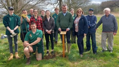 A group of 13 people posing for a photo whilst planting a tree.